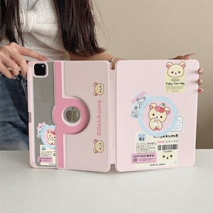 May include: Pink Rilakkuma iPad case with a bear design. The case has a magnetic closure and a stand function. The case has a clear back and a pink front cover with the text "Rilakkuma" and "Enjoy Your Day".