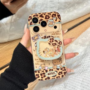 May include: A clear phone case with a leopard print design and a cartoon character. The case has a pop-up grip with a glittery interior. The case has the text "Kitty" and "OWOCASE".