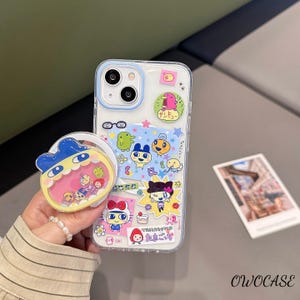 May include: A clear phone case with a blue border, decorated with colorful cartoon characters and Japanese text. A matching phone grip is attached. The case and grip feature a variety of cute, whimsical designs.