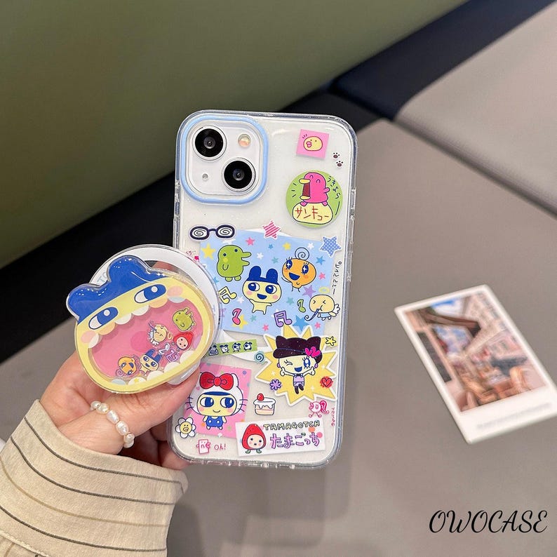 May include: A clear phone case with a blue border, decorated with colorful cartoon characters and Japanese text. A matching phone grip is attached. The case and grip feature a variety of cute, whimsical designs.