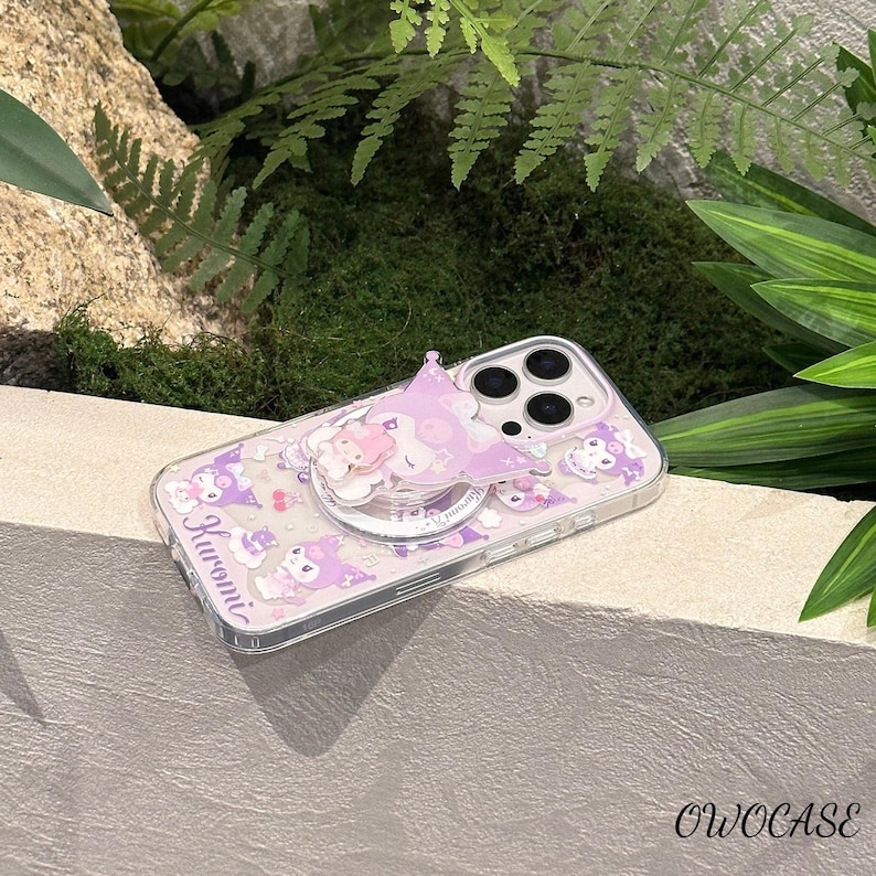 Puede incluir: Funda transparente para tel&eacute;fono con dise&ntilde;o de Kuromi morado. La funda presenta un agarre emergente con dise&ntilde;o de estrella y una peque&ntilde;a figura de Kuromi. La funda tiene la palabra "Kuromi" impresa. La marca "OWOCASE" es visible.