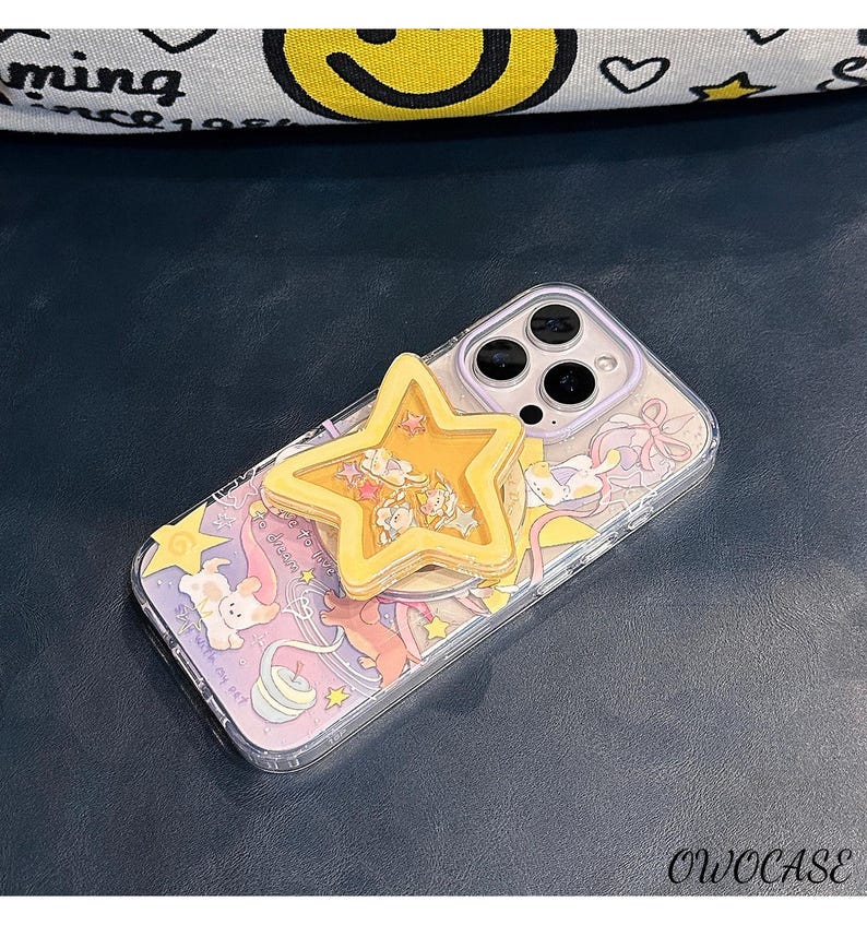 May include: A clear phone case with a colourful cartoon design and a yellow star-shaped phone grip. The case features a whimsical illustration with pastel colours and the text "dream". The phone case is designed to protect the phone while adding a decorative touch.