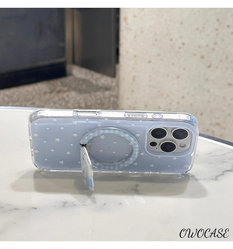May include: A clear phone case with a light blue background and white star pattern. The case has a built-in ring holder with a gingham pattern and a kickstand. The phone case has the text "OWOCASE" at the bottom.