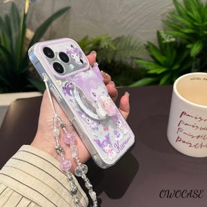 Puede incluir: Funda transparente para tel&eacute;fono con un dise&ntilde;o de personaje de dibujos animados morado y blanco. La funda tiene un soporte de anillo y una correa de cuentas con detalles en rosa y plata. La palabra "Kuromi" est&aacute; impresa en la funda.