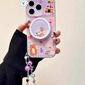 May include: A clear phone case with a lavender border and a whimsical design. The case features a shaker element with miniature desserts and bears, along with attached charms. The case has the text "OWOCASE".
