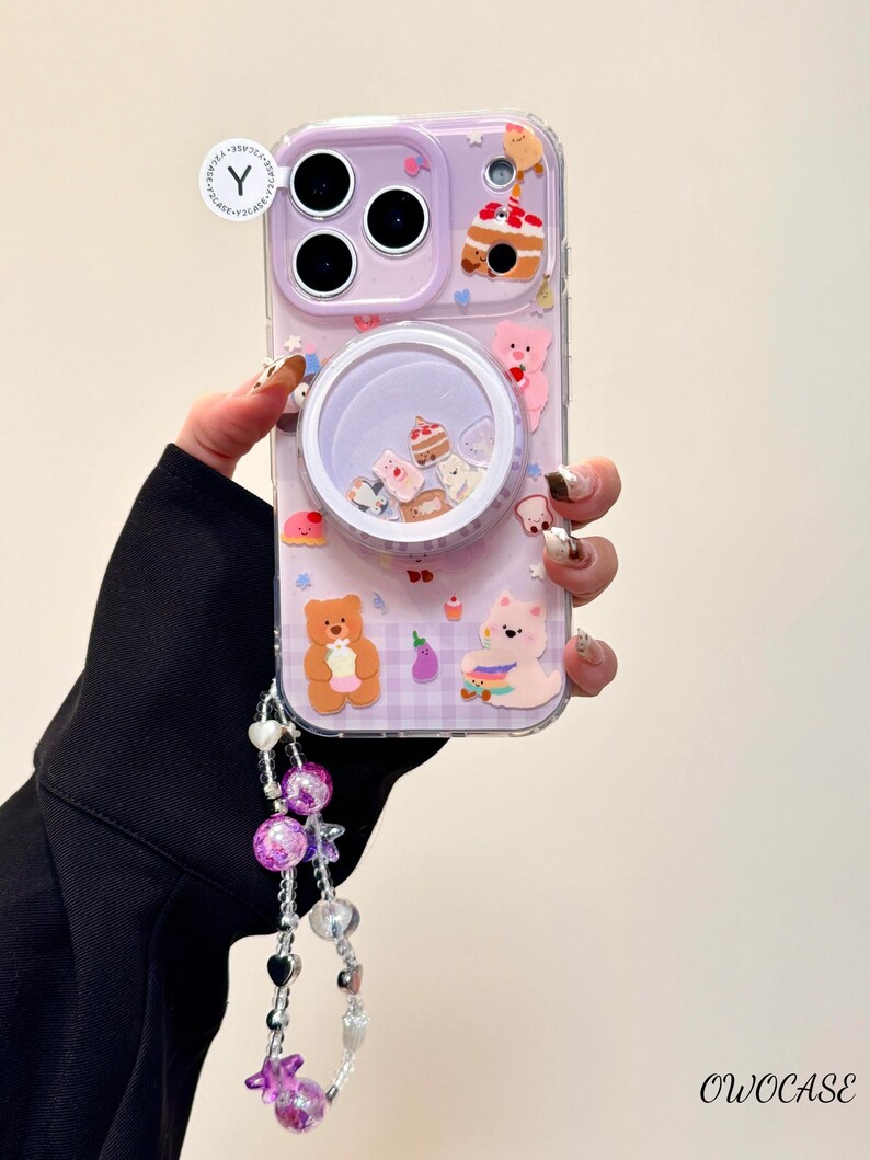 May include: A clear phone case with a lavender border and a whimsical design. The case features a shaker element with miniature desserts and bears, along with attached charms. The case has the text "OWOCASE".