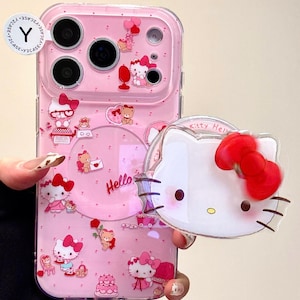 May include: Pink phone case with a clear overlay, featuring Hello Kitty and teddy bear graphics. The case has a clear, circular design with the word "Hello" in pink. A separate Hello Kitty charm is held in front.