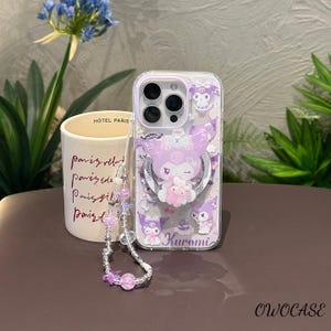 Puede incluir: Funda transparente para tel&eacute;fono con dise&ntilde;o de Kuromi morado, soporte de anillo plateado y correa de mu&ntilde;eca con cuentas. La funda presenta un personaje de dibujos animados y la palabra "Kuromi".