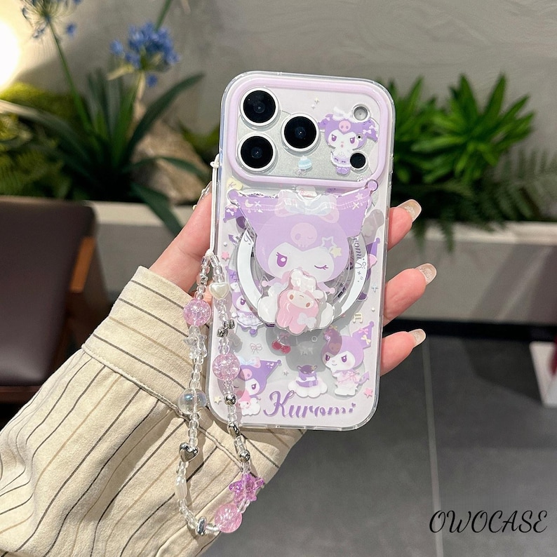 Puede incluir: Una funda transparente para tel&eacute;fono con un dise&ntilde;o de dibujos animados morado y blanco con el personaje Kuromi. La funda tiene un soporte de anillo y una correa de mu&ntilde;eca con cuentas rosas y transparentes. La funda tiene el texto "Kuromi".