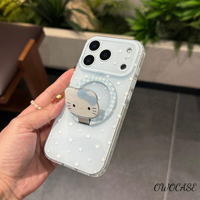 May include: A light blue phone case with a star pattern and a Hello Kitty phone grip. The case has a clear border and a circular grip with a blue and white checkered design. The Hello Kitty grip is white with a blue bow.