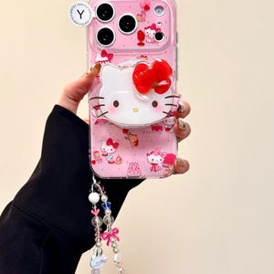 Pu&ograve; includere: Una custodia per telefono rosa con un design di Hello Kitty e un grande fiocco rosso. La custodia ha uno strato protettivo trasparente e un cinturino da polso con perline rosa e bianche. La custodia ha uno strato protettivo trasparente e un cinturino da polso.