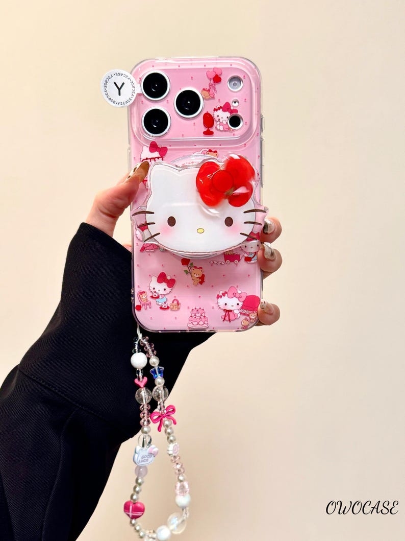 Pu&ograve; includere: Una custodia per telefono rosa con un design di Hello Kitty e un grande fiocco rosso. La custodia ha uno strato protettivo trasparente e un cinturino da polso con perline rosa e bianche. La custodia ha uno strato protettivo trasparente e un cinturino da polso.