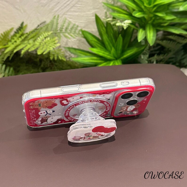 May include: A clear phone case with a red border featuring a Hello Kitty design. The case is paired with a matching pop socket. The pop socket and case have a clear background with Hello Kitty characters and red accents.