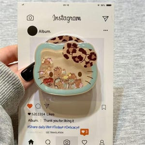 May include: A Hello Kitty-shaped phone grip with a clear, liquid-filled center containing small, colorful charms. The grip has a light blue border and a brown leopard print bow. The Instagram-style packaging includes the text "Instagram" and "Album."