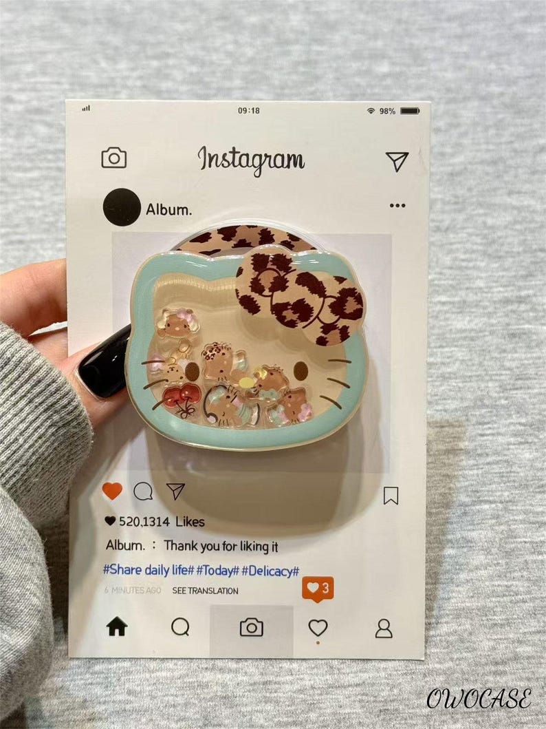 May include: A Hello Kitty-shaped phone grip with a clear, liquid-filled center containing small, colorful charms. The grip has a light blue border and a brown leopard print bow. The Instagram-style packaging includes the text "Instagram" and "Album."