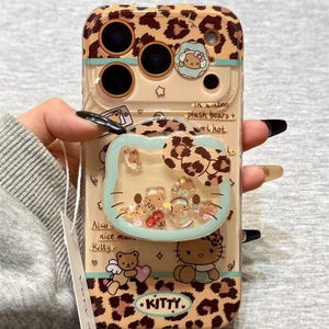 May include: A clear phone case with a leopard print design and a Hello Kitty theme. The case features a liquid-filled Hello Kitty-shaped charm with floating glitter and small figures. The text "KITTY" is visible.