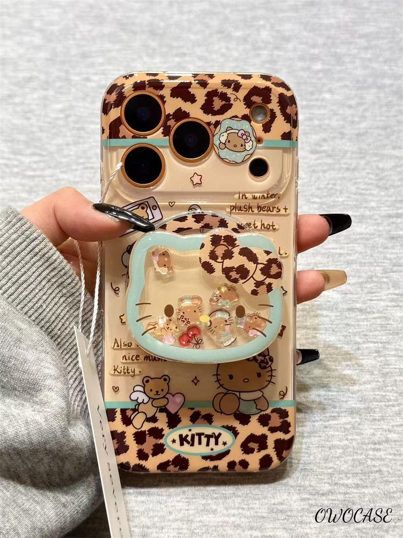 May include: A clear phone case with a leopard print design and a Hello Kitty theme. The case features a liquid-filled Hello Kitty-shaped charm with floating glitter and small figures. The text "KITTY" is visible.