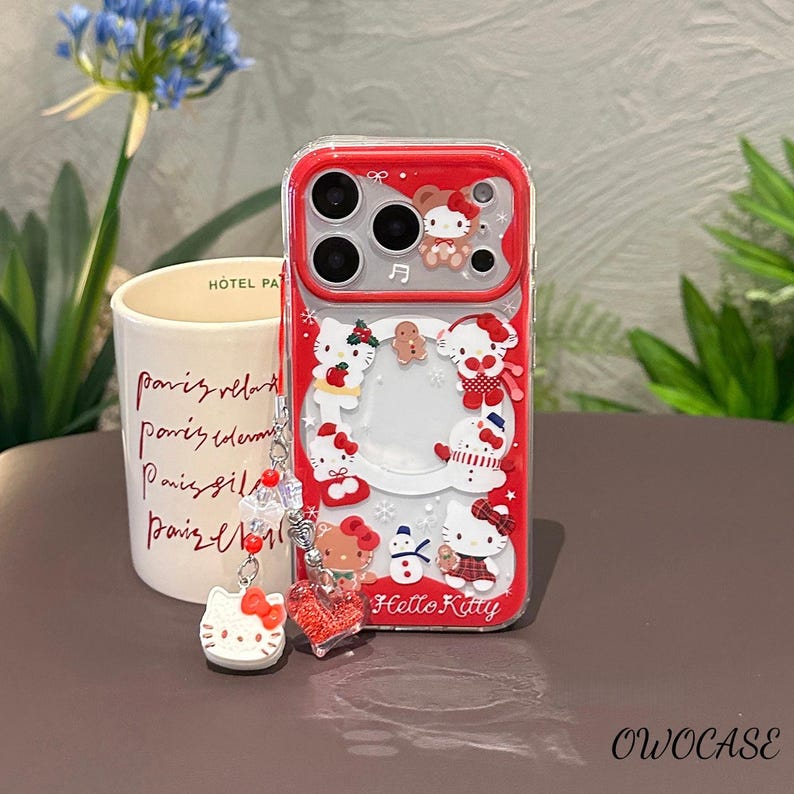 May include: A clear phone case with a red border featuring Hello Kitty designs. The case has a decorative charm with a Hello Kitty figure and a heart-shaped charm. The text "Hello Kitty" is visible on the case.