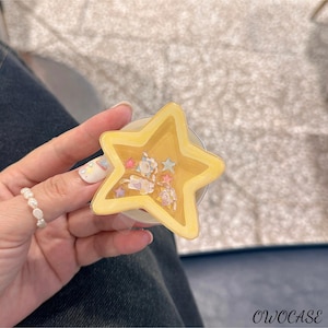 May include: A yellow star-shaped phone grip with a clear base. The grip has a glittery interior with small cartoon figures and stars. The text "OWOCASE" is visible in the bottom right corner.