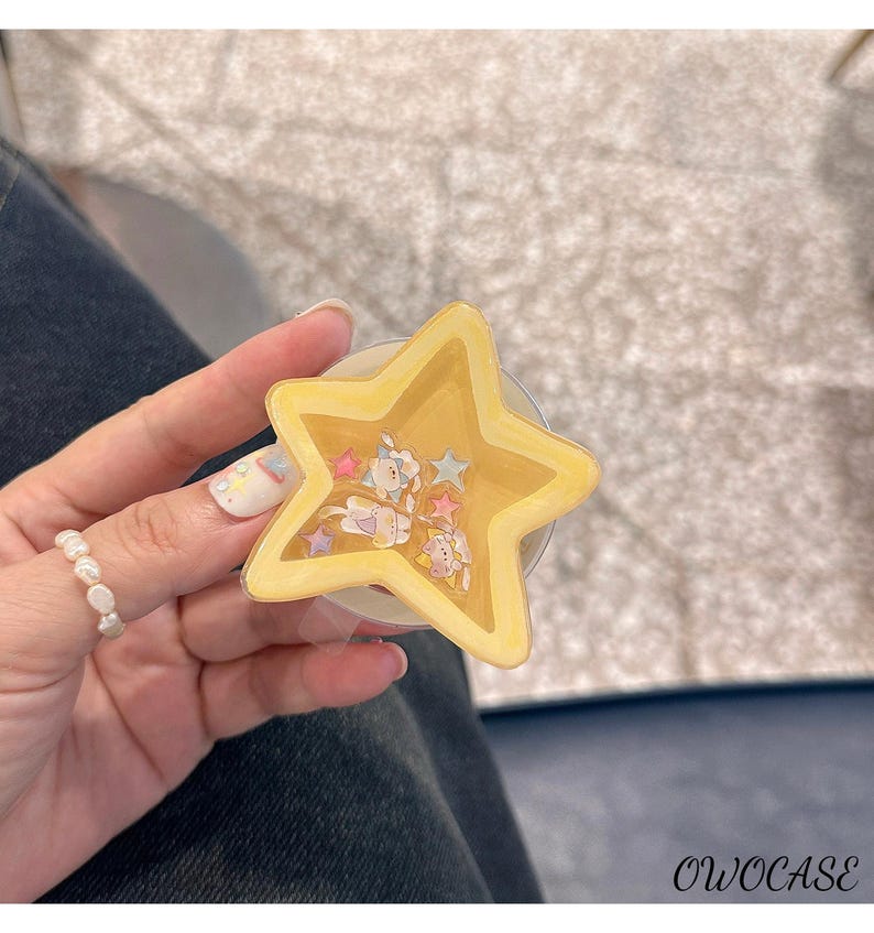 May include: A yellow star-shaped phone grip with a clear base. The grip has a glittery interior with small cartoon figures and stars. The text "OWOCASE" is visible in the bottom right corner.