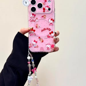 Pu&ograve; includere: Custodia per telefono rosa con disegni di Hello Kitty e cinturino da polso con perline. La custodia presenta diverse illustrazioni di Hello Kitty e le parole "Hello Kitty" in un design circolare. Il cinturino da polso include perline rosa, bianche e trasparenti e un fiocco rosa.