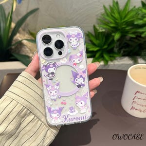 Puede incluir: Funda transparente para tel&eacute;fono con borde lila y dise&ntilde;o de personajes de dibujos animados. La funda tiene un recorte circular para la c&aacute;mara y un dise&ntilde;o impreso del personaje Kuromi. La palabra "Kuromi" est&aacute; impresa en cursiva en la parte inferior de la funda.