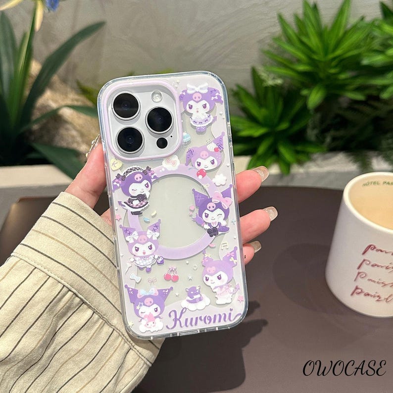 Puede incluir: Funda transparente para tel&eacute;fono con borde lila y dise&ntilde;o de personajes de dibujos animados. La funda tiene un recorte circular para la c&aacute;mara y un dise&ntilde;o impreso del personaje Kuromi. La palabra "Kuromi" est&aacute; impresa en cursiva en la parte inferior de la funda.