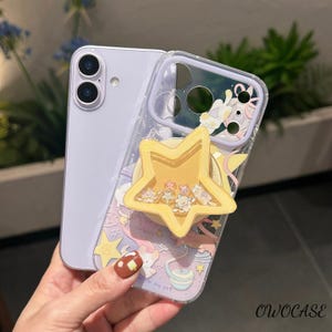 May include: A clear phone case with a yellow star-shaped pop-up grip. The case features a colourful design with stars, a unicorn, and the text "dream". The phone is a light purple colour.