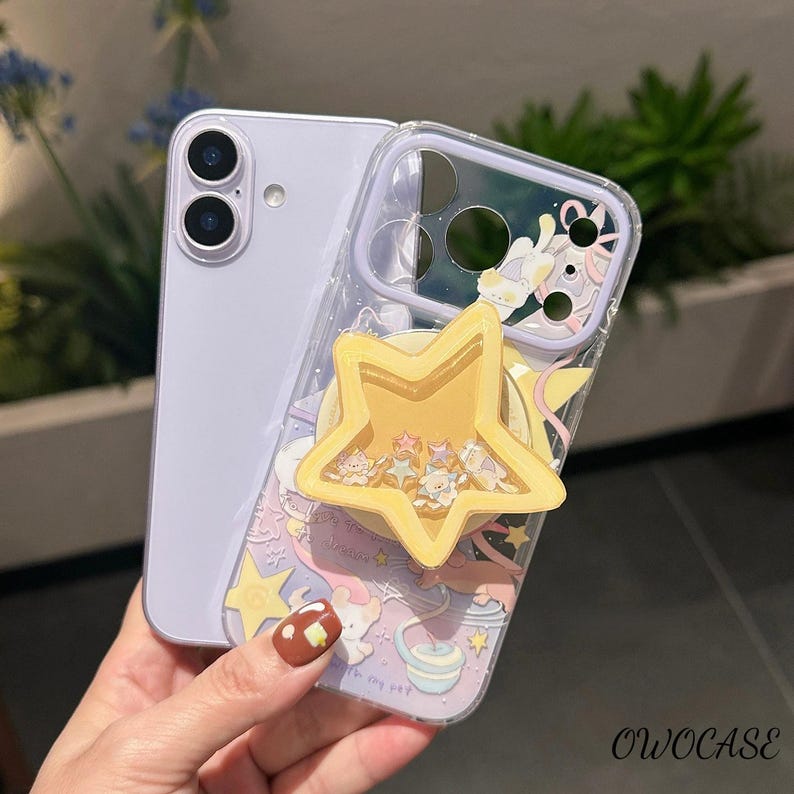 May include: A clear phone case with a yellow star-shaped pop-up grip. The case features a colourful design with stars, a unicorn, and the text "dream". The phone is a light purple colour.
