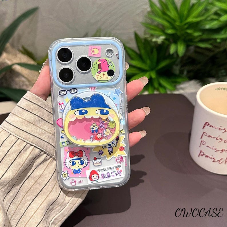 May include: Clear phone case with a blue border and colorful cartoon stickers. The stickers include a character with a blue hat and yellow mouth, a Hello Kitty sticker, and Japanese text. The case has a camera cutout and the text "OWOCASE".
