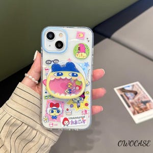 May include: Clear phone case with a blue border, decorated with colorful cartoon stickers. The stickers feature various characters and Japanese text. The case is held in a hand, with a pearl bracelet visible.