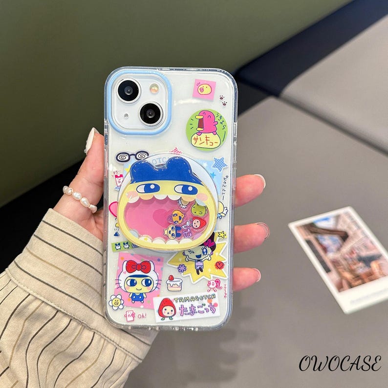 May include: Clear phone case with a blue border, decorated with colorful cartoon stickers. The stickers feature various characters and Japanese text. The case is held in a hand, with a pearl bracelet visible.