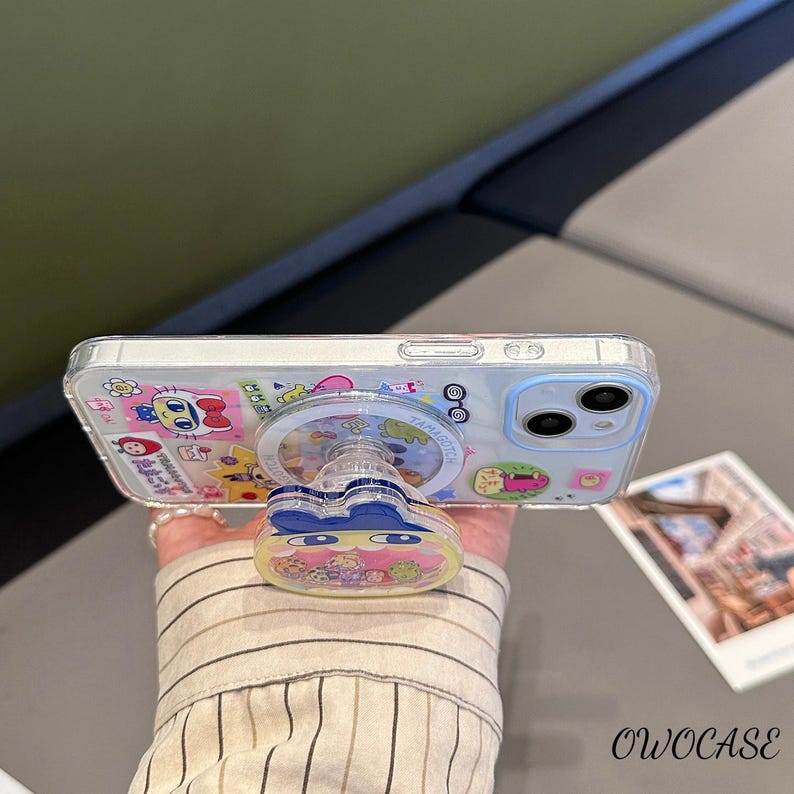 May include: Clear phone case with a cartoon design and a matching pop-up grip. The case features colorful illustrations of characters and text. The pop-up grip has a blue bow design and a clear center with more cartoon characters.
