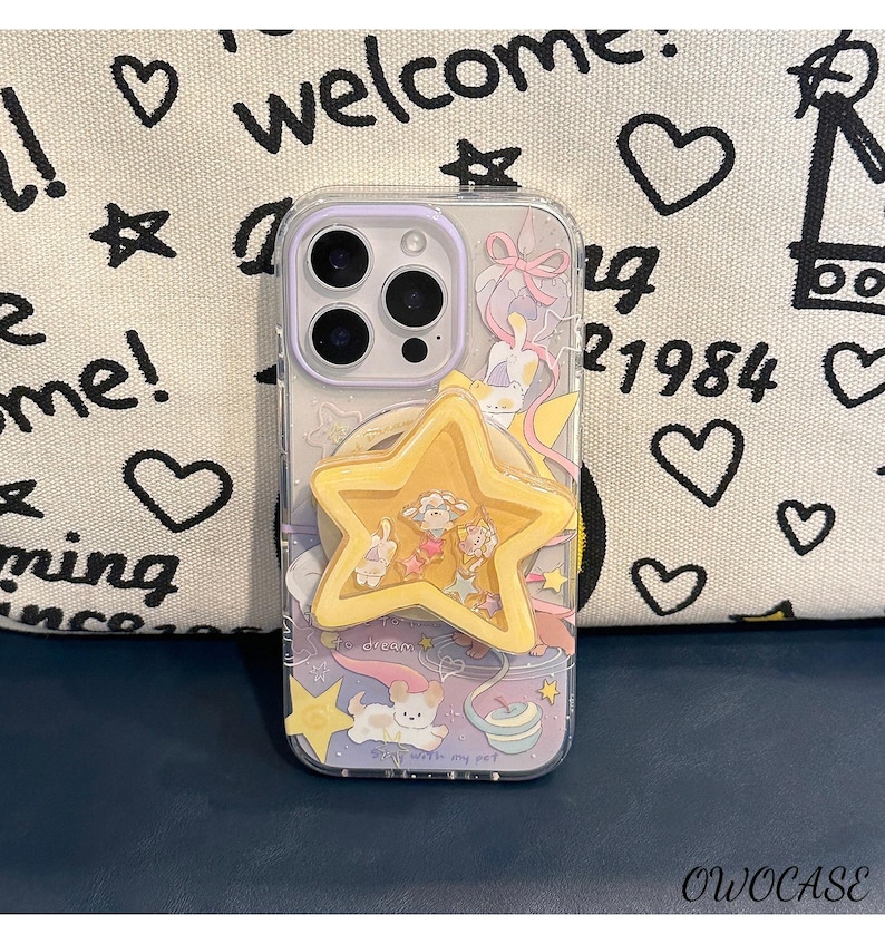 May include: A clear phone case with a playful design, including cartoon characters, stars, and a yellow star-shaped phone grip. The case has a purple border and the text "welcome" and "me!" on the background.