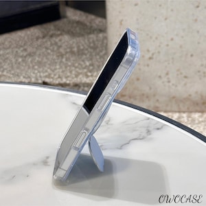 May include: Clear phone case with a built-in light blue kickstand. The case is transparent, allowing the phone's color to show through. The brand name "OWOCASE" is printed in black at the bottom right corner. The phone is resting on a white marble surface.