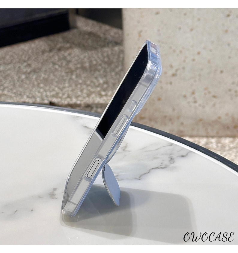 May include: Clear phone case with a built-in light blue kickstand. The case is transparent, allowing the phone's color to show through. The brand name "OWOCASE" is printed in black at the bottom right corner. The phone is resting on a white marble surface.
