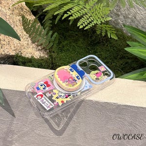 May include: Clear phone case with colorful cartoon character stickers and a pop-up grip. The grip features a pink heart-shaped design with a blue character. The case has a light blue border and is decorated with various stickers.