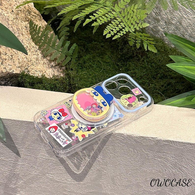 May include: Clear phone case with colorful cartoon character stickers and a pop-up grip. The grip features a pink heart-shaped design with a blue character. The case has a light blue border and is decorated with various stickers.