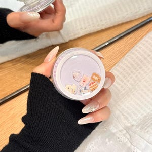 May include: A clear, round phone grip with a light purple interior filled with small, colorful food-themed charms. The grip is held in a hand with long, manicured nails. The brand name "OWOCASE" is visible in the bottom right corner.