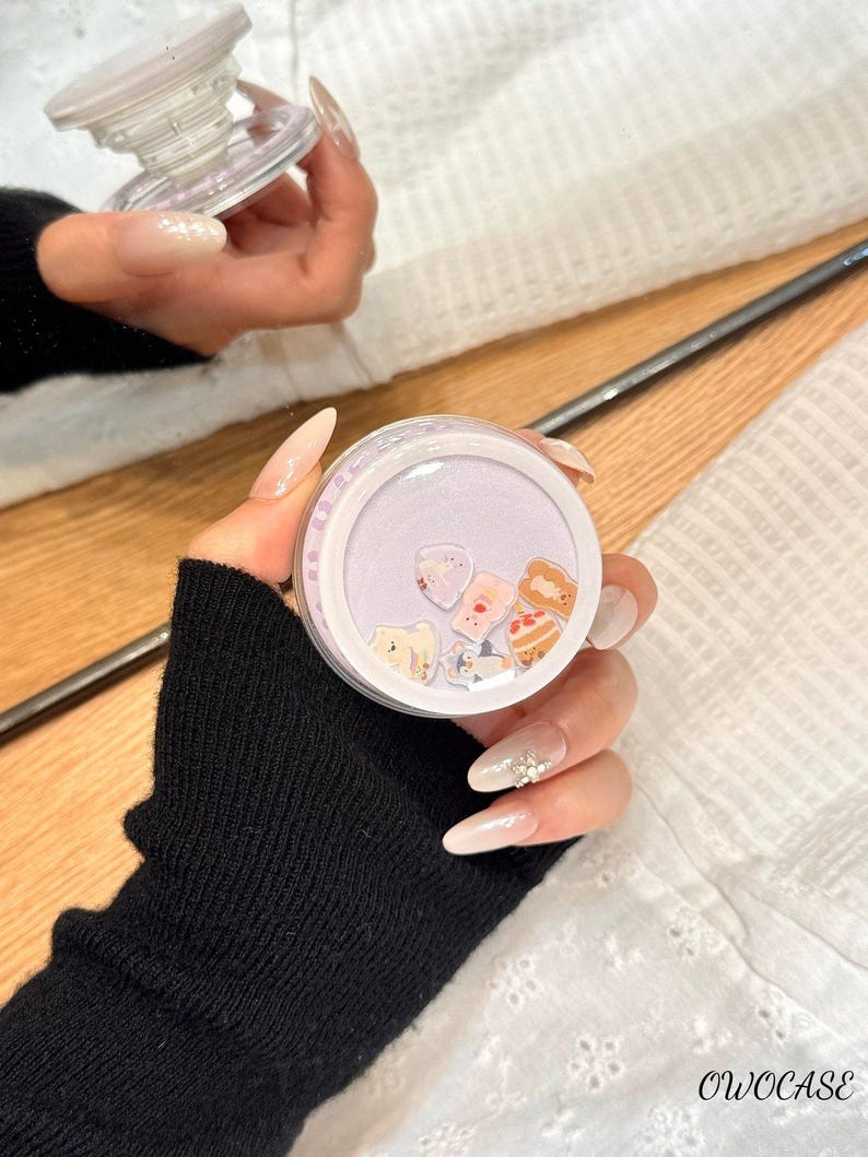 May include: A clear, round phone grip with a light purple interior filled with small, colorful food-themed charms. The grip is held in a hand with long, manicured nails. The brand name "OWOCASE" is visible in the bottom right corner.