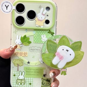 Op de afbeelding: Een lichtgroene telefoonhoes met een cartoon konijn ontwerp. De hoes heeft verschillende konijn illustraties, harten en de tekst "happiness" en "Everything goes well Good luck always accompanies you". Een konijnvormige popsocket is bevestigd.