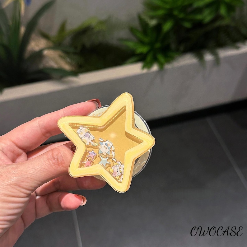 May include: A yellow star-shaped phone grip with a clear centre filled with small star and character-shaped decorations. The phone accessory has a white base and is held in a hand. The text "OWOCASE" is visible in the bottom right corner.