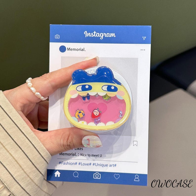 May include: A blue and yellow phone grip with a cartoon character design. The grip features a pink interior with small character illustrations and the Instagram logo. The text on the packaging includes "Memorial." and "OWOCASE."