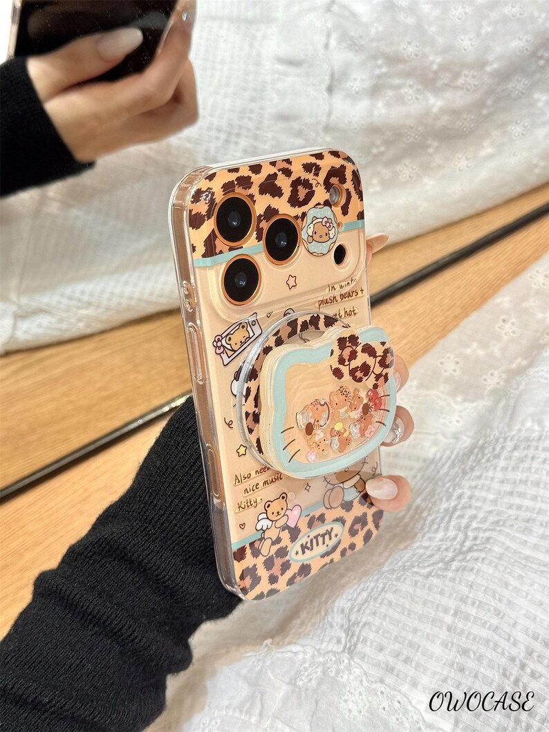 May include: A clear phone case with a leopard print design and a pop-up grip. The case features illustrations of bears and the word "KITTY." The text "Also need nice music Kitty" is also visible. The case is held by a person.