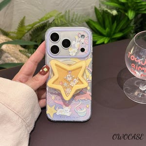 May include: Clear phone case with a whimsical design featuring a yellow star-shaped pop socket. The case has a lilac border and a colourful illustration with a cat, stars, and the text "Stay with my cat."