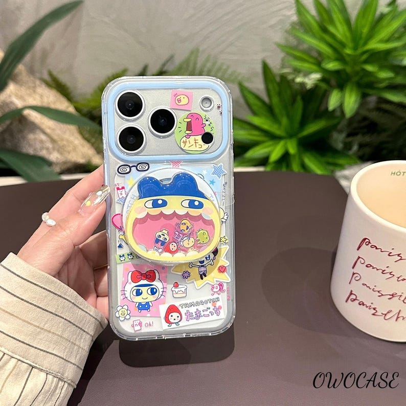 May include: Clear phone case with a blue border and a cartoon design. The case features a pop-up character with a large mouth and various other cartoon characters. The case also has a pink bow and the text "Tamagotchi".