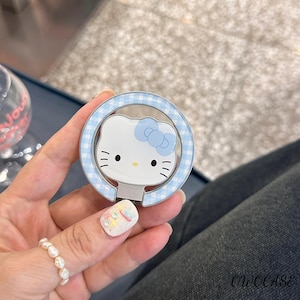 May include: A white and blue phone ring holder featuring a cartoon cat face with a blue bow. The ring holder has a light blue gingham pattern around the cat's face. The base is silver and has the text "OWOCASE".