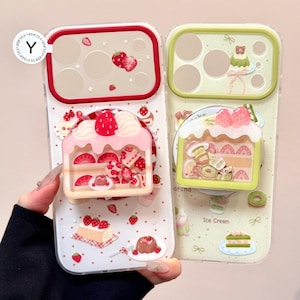 以下が含まれることがあります： デザートをテーマにした2つの携帯電話ケース。1つのケースはイチゴケーキ、もう1つはキウイのスライスが入ったケーキが特徴です。どちらのケースも、赤と緑のアクセントが付いた透明な背景です。ケースはイチゴやその他のデザート要素で飾られています。