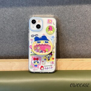 May include: Clear phone case with a colorful cartoon design. The case features a blue and yellow character with a pink mouth, surrounded by other cartoon characters and Japanese text. The case has a blue camera ring and a clear back.