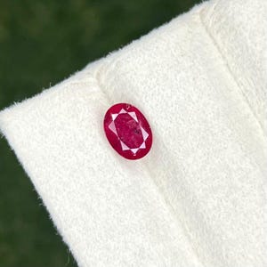Può includere: Una gemma di rubino taglio ovale con un colore rosso intenso e una superficie sfaccettata. La pietra mostra una struttura interna chiara con inclusioni naturali visibili e una finitura lucida che riflette la luce sulla sua superficie.